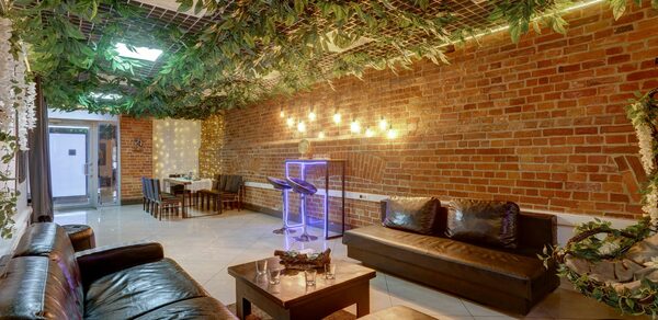 Лофт Семеновский. Лофт-пространство с кирпичными стенами и подвесным потолком, украшенным зеленью. Кожаные диваны и деревянный стол в центре. В углу стол с креслами. Светильники на стене. Яркая неоновая установка. Пол плиточный.