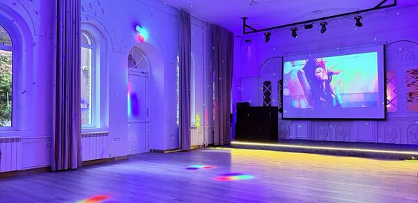 Лофт Софт. Просторное помещение со светлым паркетным полом и фиолетовой подсветкой. На стене большой экран с проектором, справа диджейский пульт. Окна с арочными рамами, закрытыми шторами. Потолочные светильники на треке и декоративные лепные элементы.