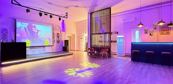 Лофт Софт. Просторное мероприятие с деревянным полом и высоким потолком, украшенным лепниной. В центре — сцена с большим экраном. Справа — барная стойка с подвесными светильниками. Красочное освещение, несколько столиков и стульев.