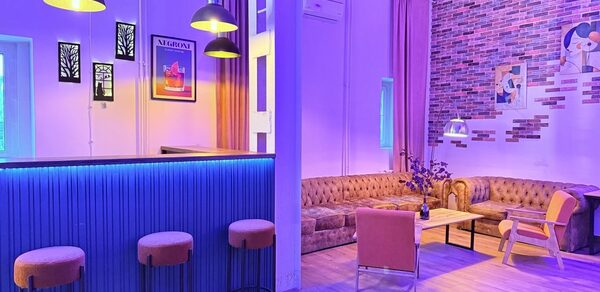 Лофт Софт. Праздничное пространство с барной стойкой, облицованной синими панелями, и четырьмя барными стульями. Левый угол оформлен в виде зоны отдыха с коричневым диваном, креслом и журнальным столиком. Стены украшены картинами и кирпичной кладкой. Освещение фиолетово-жёлтое.