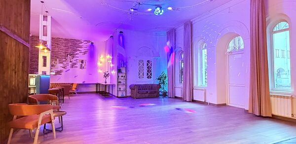 Лофт Софт. Просторное помещение с деревянным полом, цветным освещением и кирпичной стеной. По краям расположены диван и стулья, большие арочные окна с занавесками. Подвесные светильники и прожекторы обеспечивают яркую подсветку.