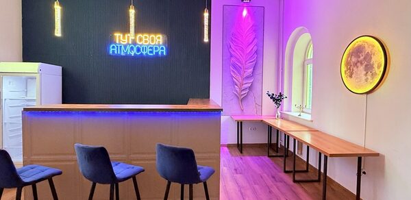 Лофт Лайф. Барная стойка с барными стульями, неоновая надпись на синей стене, подвесные светильники, декоративная панель с подсветкой, деревянные столы вдоль стены, окна с арками, деревянный пол, декоративный круг с подсветкой на стене.