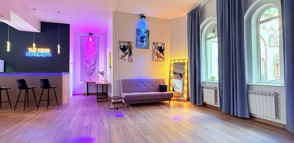 Лофт Лайф. Просторная гостиная с деревянным полом, барной стойкой с тремя черными стульями слева. На стене вывеска с подсветкой. Большое зеркало в золотой раме, диван у стены и картины с подсветкой. Окна с высокими шторами, яркое разноцветное освещение.