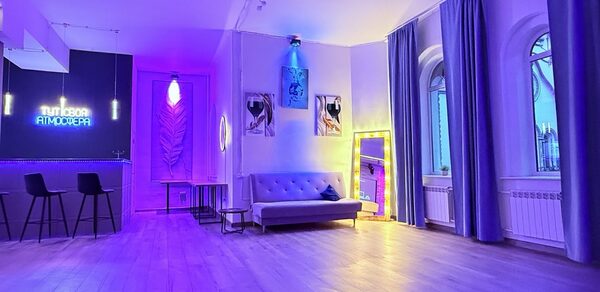 Лофт Лайф. Современное помещение с деревянным полом, высокой потолочной лампой и фиолетово-синим неоновым освещением. Зона с барной стойкой и стульями, диван, большие арочные окна с шторами, декоративные картины, высокое зеркало с подсветкой.