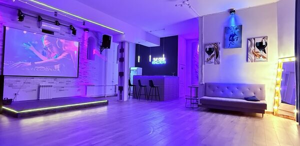 Лофт Лайф. Просторное помещение с деревянным полом, сцена с проектором и подсветкой, барная стойка с двумя стульями, диван у стены, подсвечен LED-лентами. На стенах картины и неоновая вывеска. Освещение разных цветов, в основном фиолетовые и синие тона.