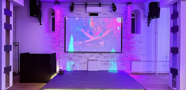 Лофт Лайф. Сцена с проектором и изображением диджея, чёрные колонки по бокам, светодиодная подсветка, паркетный пол, белые стены с декоративной кирпичной кладкой, потолочные светильники, радиаторы отопления, фиолетовое освещение.