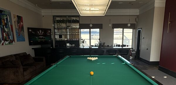 Студия Н. Бильярдный зал с большим зелёным столом в центре. Барная стойка с высокими стульями вдоль стены. Кожаный диван слева. Подвесное освещение и настенные картины. Коричневый пол, светлые стены с тёмными акцентами.