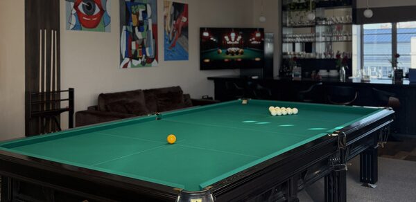 Студия Н. Бильярдная комната с столом для русской пирамиды, рядом с ним диван и телевизор, на стенах картины в ярких цветах. Стойка с напитками и бокалами, барные стулья, окно с видом на город, светлая перегородка с освещением.