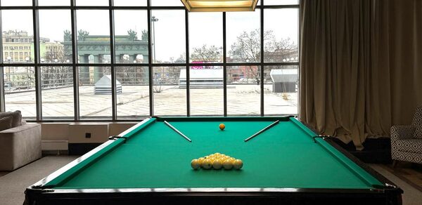 Студия Н. Бильярдный стол с зелёным сукном, пара киев, шары треугольником. Просторное помещение с большими окнами, тип штора кремового цвета справа. Потолочный светильник над столом. Слева диван и кресло с узором. Панорамный вид на городские крыши.