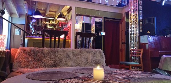 Лофт арт-хаус с террасой площадью 65 м². Интерьерное пространство с двухъярусной планировкой. В центре мягкая зона с диваном и столом, на котором стоит горящая свеча. На заднем плане видна барная стойка с полками для бутылок. Освещение из подвесных ламп и гирлянд.