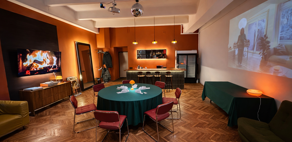 The Gallery Loft (Лофт Галерея) . Просторная комната с деревянным паркетом, круглым столом с цветами и красными стульями. Встроенный проектор, барная стойка с высокими стульями, черный холодильник. Оранжевые стены с изображением камина, потолочные прожекторы и зеркальный шар.