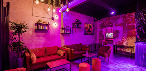 Дизайнерский лофт с караоке и активностями возле метро. Лофт-пространство с кирпичными стенами, диванами и креслами красного цвета, низкими столиками, полками с декором. Освещение фиолетовое и теплое. На стенах растительные декорации и картины. Телевизор у стены, декоративные лампы на потолке.