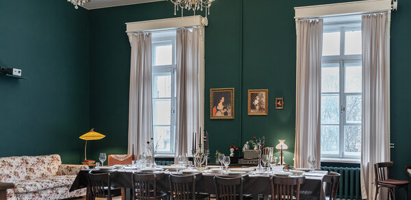 Пространство Имена. Винтажный лофт . Винтажная гостиная с зелеными стенами, двумя высокими окнами с белыми шторами. Просторный стол с черной скатертью, сервированный на двенадцать человек. Деревянные стулья, диван с цветочным узором. Люстры с лампами, картины на стене.