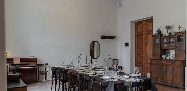 Пространство Имена. Винтажный лофт . Большая столовая с длинным деревянным столом и стульями, накрытыми черной тканью, сервировка посуды и бокалов. Слева пианино, шкаф и зеркало на стене. Деревянный пол, стены светлые. Потолочная люстра с подвесками.