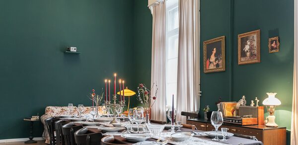 Пространство Имена. Винтажный лофт . Столовая с длинным накрытым столом на двенадцать персон, деревянные стулья, плотные белые шторы, зеленые стены, картины в рамках. На комоде лампы и декор, в центре стола свечи и цветы. Потолочная люстра, паркетный пол.