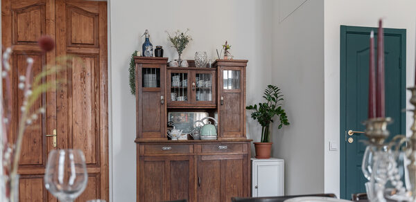 Пространство Имена. Винтажный лофт . Столовая с деревянным буфетом, на котором стоят вазы и растения. Перед буфетом накрытый стол с белыми тарелками и бокалами. Крупные деревянные двери справа закрыты, рядом зеленая дверь, рядом с буфетом комнатное растение.