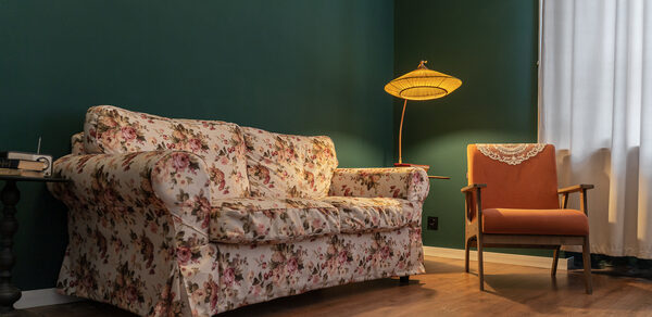 Пространство Имена. Винтажный лофт . Гостиная с темно-зелеными стенами и деревянным полом. Диван с цветочной обивкой и оранжевое кресло. Напольная лампа с тканевым абажуром, белая занавеска на окне. Маленький круглый стол рядом с диваном.