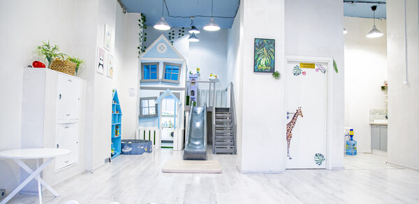 Лофт Облака. Зал Баунти на Полярной. Пространство детской игровой с зоной для занятий и горкой, большая белая комната с высоким потолком, белые стены и пол. Голубая игровая структура в виде домика с лестницей и горкой. Яркие акценты: жираф на двери, растения в горшках. Освещение подвесное.
