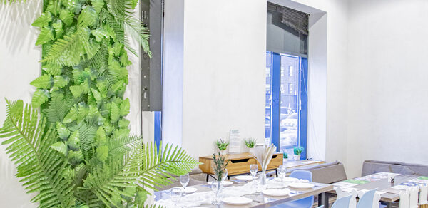 Лофт Облака. Зал Баунти на Полярной. Современное помещение с белыми стенами, окнами, столом с белыми тарелками и бокалами. Голубые стулья, светлое деревянное напольное покрытие, декоративные растения на стене и столе, черный диван у стены, естественное освещение.