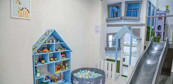 Лофт Облака. Зал Баунти на Полярной. Детская игровая зона с горкой, стеной в виде дома и игрушечным домиком. Голубой стеллаж с игрушками и круглым бассейном с мягкими шарами. Светлые стены, частичное зелёное растение на стене, картина с тигром.