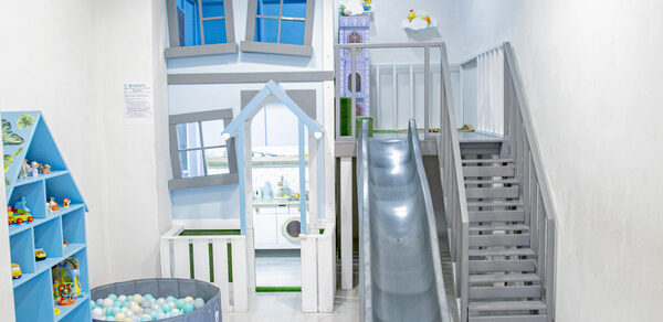 Лофт Облака. Зал Баунти на Полярной. Детская игровая зона с деревянным домиком, лестницей и горкой. Стены и пол светлые, корзина с цветными шарами. Шкаф с игрушками слева, домик окрашен в голубые и белые тона. Яркое освещение, на стене заметен текст.
