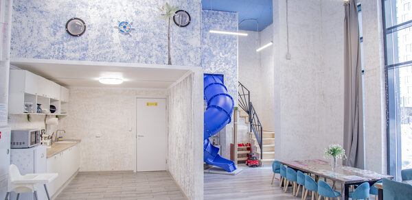 Лофт Облака. Зал Баунти на Сельскохозяйственной. Открытое пространство с высокими потолками, игровой горкой, зоной кухни и длинным столом с мягкими бирюзовыми стульями. Стены белые с синим узором, полы светлые. Естественное и искусственное освещение, большие окна, деревянная лестница.