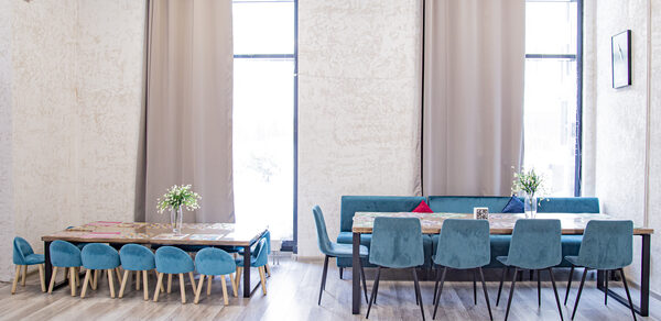 Лофт Облака. Зал Баунти на Сельскохозяйственной. Просторное помещение с двумя длинными столами и стульями с мягкой обивкой бирюзового цвета. Высокие окна с бежевыми шторами. Светлые деревянные полы. На столах цветочные композиции. Стены светлые с текстурой.