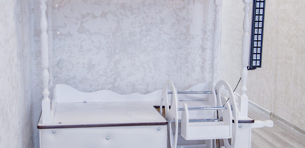 Лофт Облака. Зал Баунти на Сельскохозяйственной. Деревянная тележка с белыми и светло-коричневыми элементами, расположена у светлой стены с текстурой. Рядом светлая поверхность пола с деревянным узором. На тележке есть полки и стилизованные колеса.