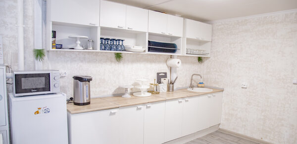 Лофт Облака. Зал Баунти на Сельскохозяйственной. Кухонное помещение с белыми шкафами и светлыми столешницами. На столе микроволновка, термопот и кухонные принадлежности. Открытые полки с посудой. Белые стены, светло-серый пол, круглые лампы на потолке. Декор из зелени.