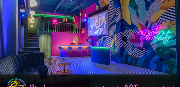 Лофт Май. Зал Мира. Караоке-зал с яркими стенами в абстрактных узорах, неоновая надпись, барная стойка с высокими стульями, черный диван, черно-белый полосатый пол, большая проекция на стене, подвесные круглые светильники, розовая барная зона под лестницей. Текст: "Профессиональное караоке AST в подарок".