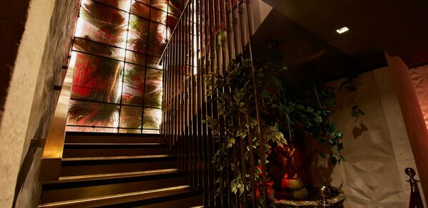 Микс Afterparty(После вечеринки). Лестница с металлическими перилами ведет вверх. Стена украшена панелями с абстрактным цветочным узором и подсветкой. Справа стоит декоративное растение в вазе. Освещение мягкое, коричнево-золотистые оттенки. Панели чередуют красные и зеленые цвета.