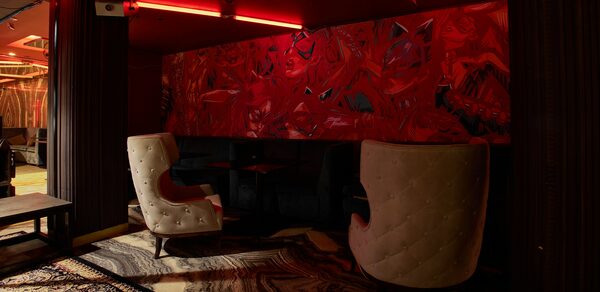 Микс Afterparty(После вечеринки). Лаунж-зона с двумя высокими белыми стульями, черным диваном, столиком. Красные абстрактные рисунки на стене. Потолочные красные светильники, ковровое покрытие с узором. Текстуры дерева на колоннах и полу.