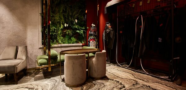 Микс Afterparty(После вечеринки). Современное помещение с зелёной стеной из растений, диваном с зелёной обивкой, круглым столом и двумя серыми креслами. Напольное покрытие из мрамора с волнистыми узорами. Красные стены, настенное освещение, справа часть гардеробной стойки.