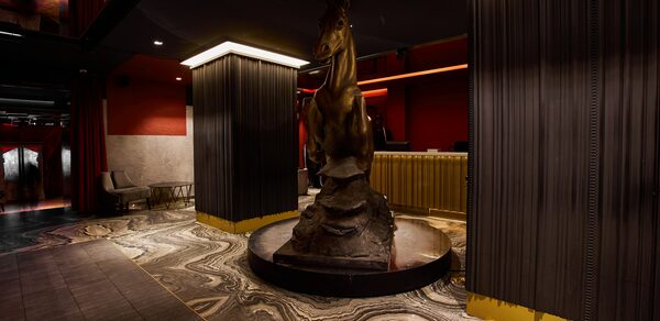 Микс Afterparty(После вечеринки). Коммерческое пространство с темными потолками, красными и бордовыми стенами. В центре статуя лошади на камне. Желтая барная стойка, мраморный пол со сложным узором. Диван в углу, приглушенное освещение, колонны с вертикальными линиями.