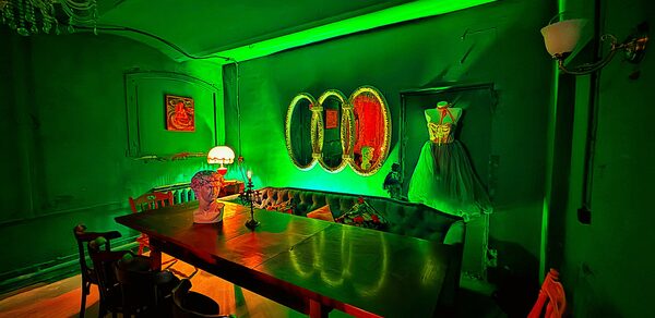 Лофт «Рич» (RICH). Зеленый интерьер с длинным деревянным столом, окруженным черными стульями. На столе лампа с абажуром. На стене три зеркала овальной формы. Угловая софа с подушками. Платье на манекене. Зеленая подсветка. Потолочный светильник.