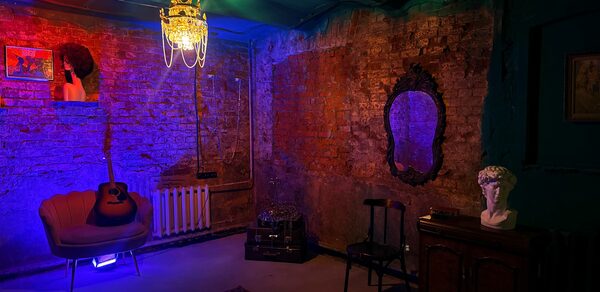 Лофт «Рич» (RICH). Интерьер с кирпичными стенами; потолочное освещение в виде люстры; мягкое кресло с гитарой; зеркало в резной раме; статуя на тумбе; ковёр на полу; разноцветная подсветка; картины на стене; табурет и радиатор вдоль стены; тёмные и теплые цвета.