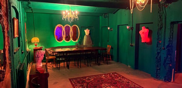 Лофт «Рич» (RICH). Комната с зелеными стенами и низким потолком. Длинный деревянный стол окружен стульями, слева бюст, на столе лампа с абажуром. Потолочные и настенные светильники. На полу яркий ковер с узорами. На стене три зеркала и розовый торс.