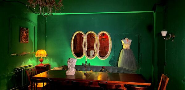 Лофт «Рич» (RICH). Зал с зелеными стенами, длинный деревянный стол с стульями, бюст на столе. Слева торшер с желтым абажуром, на стене картина. Справа платье на манекене. Тройное овальное зеркало на стене. Подвесная люстра.