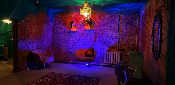 Лофт «Рич» (RICH). Кирпичные стены, два кожаных кресла, кресло с гитарой, потолочная люстра, разноцветное освещение, радиатор, ковер с узором, зеркало на стене, бюст на столике, бетонный пол.