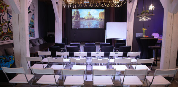 Сатурн Лофт. Многофункциональное помещение с деревянными балками, экраном и проектором. Зал оформлен стульями в несколько рядов, на стене вывешен белый экран. Имеются черные стулья, белая доска и высокий стол с цветами. Потолок украшен гирляндами и светильниками.