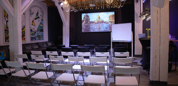 Сатурн Лофт. Современное лофт-пространство с деревянными балками и потолком, оборудованное белыми складными стульями, проектором с изображением города, экраном, флипчартом. На стенах картины, гирлянды лампочек, зона с диваном, черные барные столы.