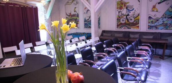 Сатурн Лофт. Пространство с рядами черных и белых стульев, черные круглые столы, белые деревянные балки, яркие картины на стенах, фиолетовая шторка, ноутбук, ваза с желтыми цветами, светильники в клетках, два граната.