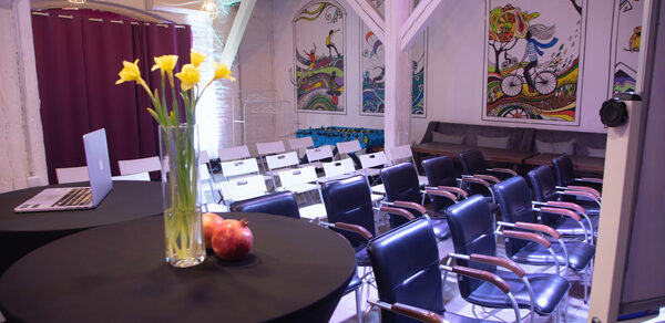 Сатурн Лофт. Пространство для мероприятий с рядами стульев и обеденными столами. Стулья черные с деревянными подлокотниками; столы покрыты черной тканью, на одном из которых ваза с желтыми цветами и яблоки. Белые стены с яркими настенными рисунками. Потолок деревянный. Освещение теплое.