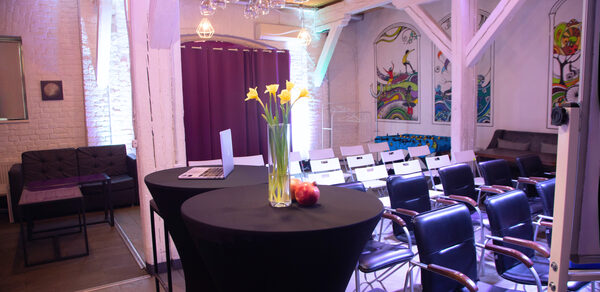 Сатурн Лофт. Лофт-пространство с белыми кирпичными стенами, деревянным потолком и балками. Ряд белых стульев, черные столы с вазой и фруктами. Настенная роспись, фиолетовые шторы. Потолочные светильники с лампочками. Черные кресла и диван.