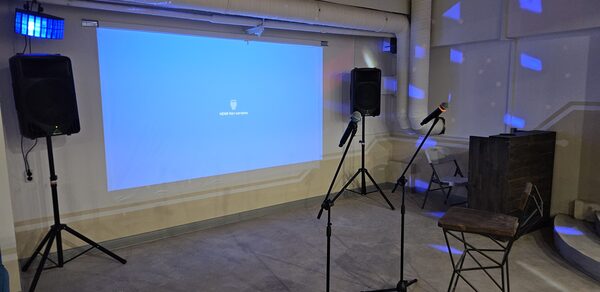 Караоке-лофт с бильярдом. Клубное пространство с проекционным экраном и двумя микрофонами на стойках. Два крупных динамика по бокам. Стул с высокой спинкой рядом с микрофонами. Серый пол, стены светлые. Подвесные трубы и вентиляция вверху. Синие световые эффекты на стенах.