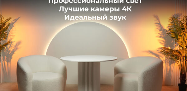 Лофт Восторг. Современное помещение с двумя белыми креслами и круглым столом. Задний план подсвечивается оранжевыми прожекторами. На стене текст: "3 локации в одном зале. Профессиональный свет. Лучшие камеры 4К. Идеальный звук." По бокам искусственные пальмы.