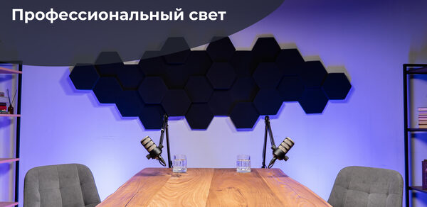 Лофт Позитив. Современное пространство с деревянным столом и двумя серыми креслами. На столе стоят микрофоны и стаканы с водой. На стене шестиугольные черные панели. Освещение фиолетовое. Слева стеллаж с книгами. Надпись: «ТОПовая техника. Профессиональный свет».