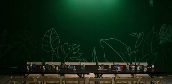 Хендмейдщики. Лофт в лесной тематике. Большой стол с рядами светлых деревянных стульев, расположенный перед зеленой стеной с изображениями листьев. На столе расставлены различные предметы. Светлый деревянный пол, точечное освещение сверху.