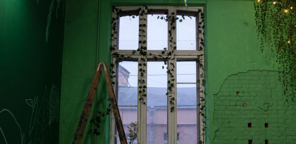 Хендмейдщики. Лофт в лесной тематике. Комната с зелеными стенами, декорированными рисунками растений, и большим окном с занавеской из искусственных листьев. У окна стол с крафтовыми материалами. Справа кирпичная стена, частично оштукатуренная. Деревянная лестница в углу.