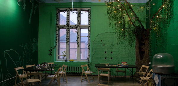 Хендмейдщики. Лофт в лесной тематике. Зеленое помещение с высоким потолком и декоративными элементами. Стены оформлены в виде деревьев с гирляндами. Несколько складных столов и деревянных стульев. Окно с белыми рамами и решеткой. На полу деревянные панели. Освещение точечными светильниками.